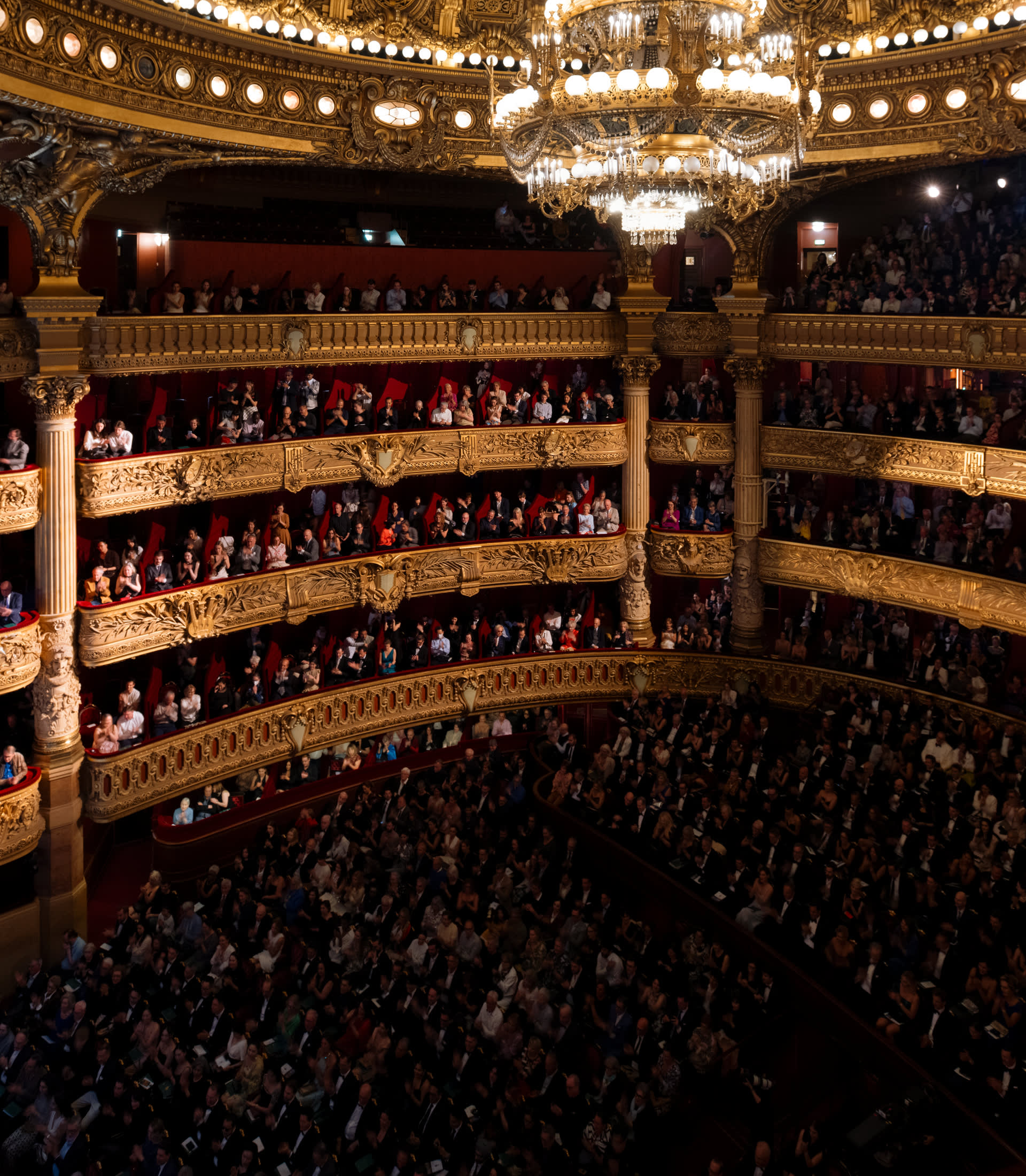 An audience seated in the ornate auditorium of the Palais Garnier opera house under a large crystal chandelier