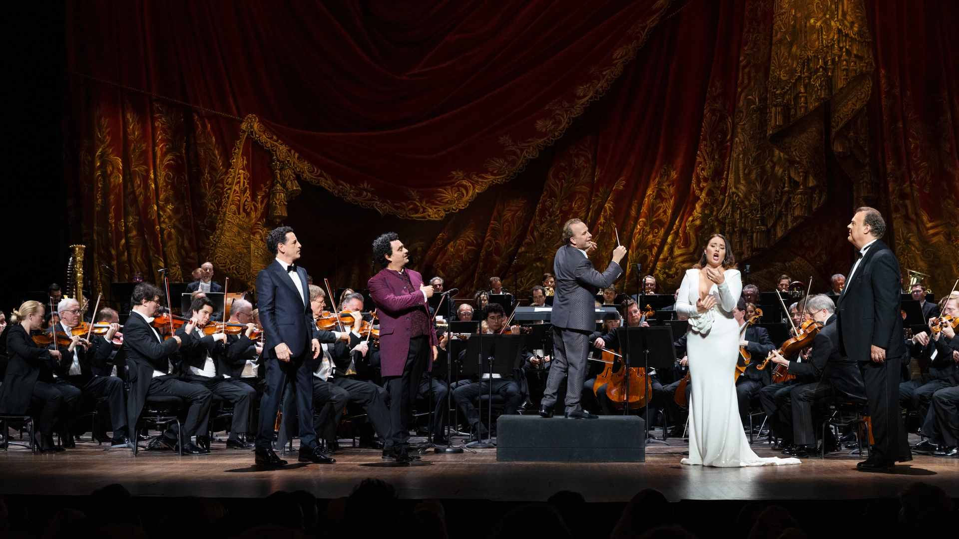 Opera performers holding hands and bowing on stage in front of an orchestra and a red curtain